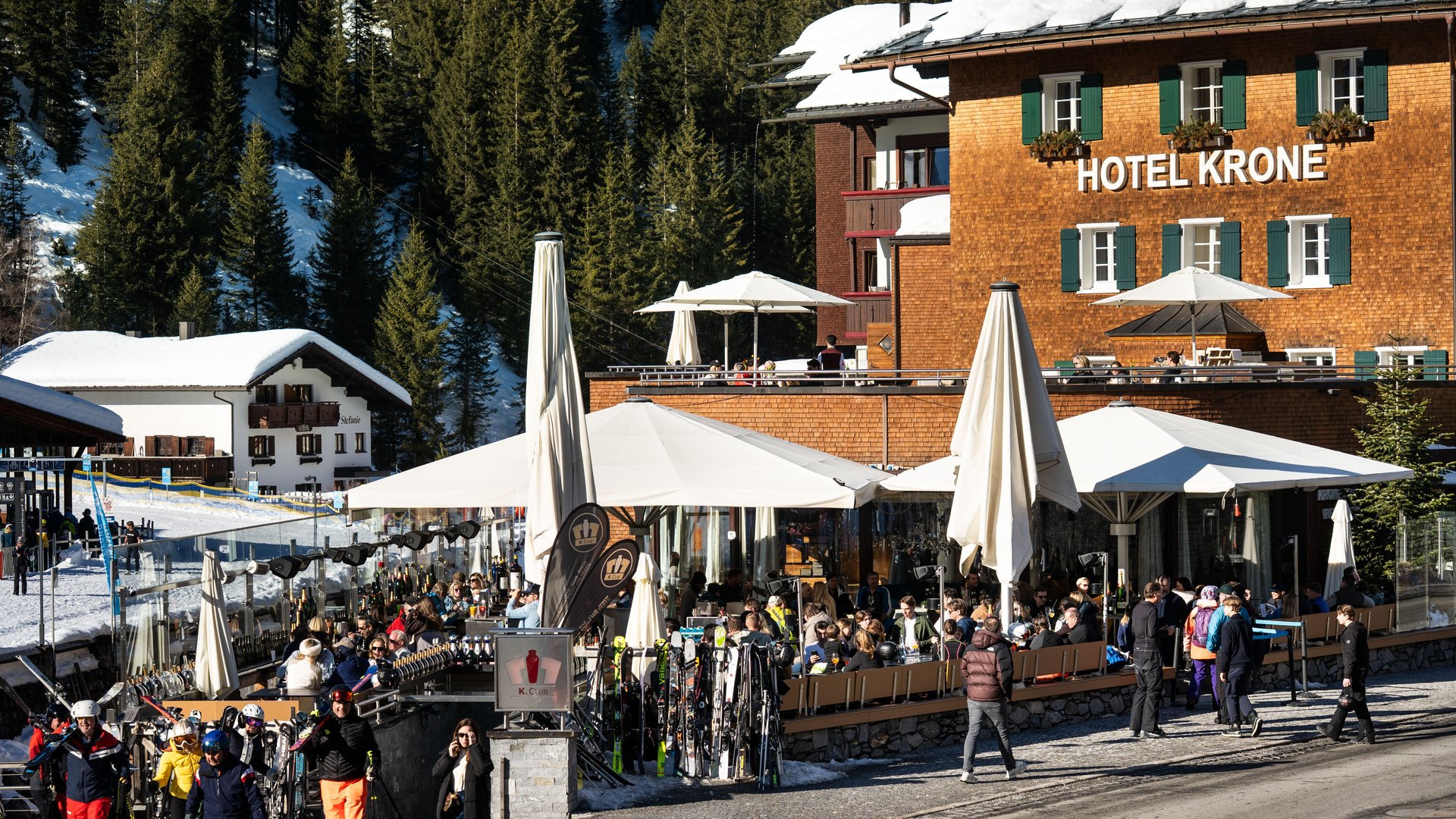 Ski Resort Erlebnisse in Lech Blick auf das Romantik Hotel “Die Krone von Lech” mit Skifahrern und Außenbereichen im Winter bei strahlendem Sonnenschein.